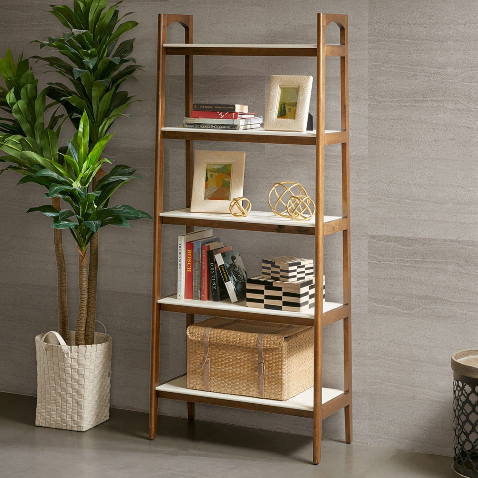 The Mid-Century Design 5 Shelf Bookcase in Off White/Pecan holds framed photos, decor, books, and baskets. A large potted plant in a woven basket stands beside it against a gray textured wall.
