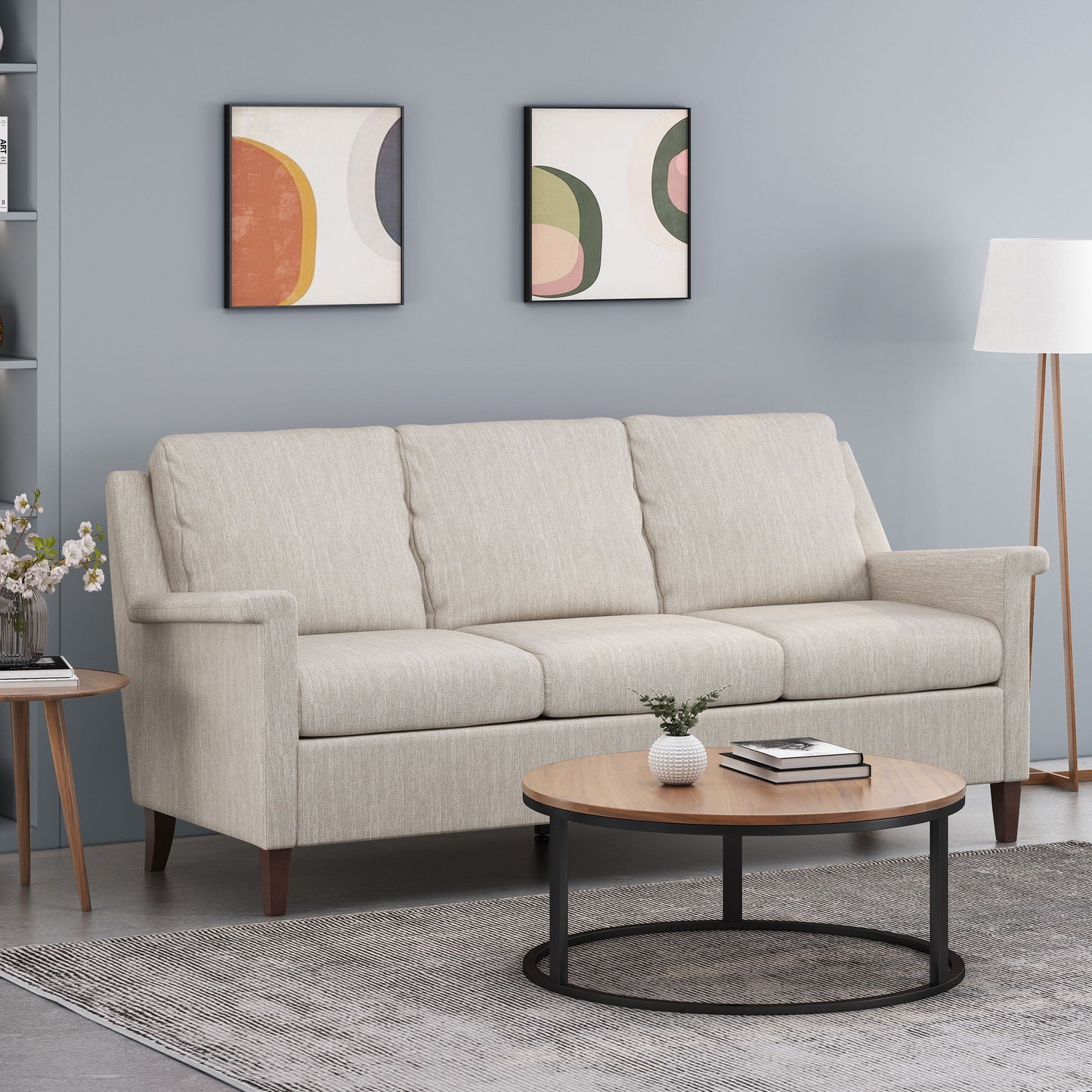 A modern living room featuring a 3 Seater Contemporary Design Sofa in light beige, round wooden coffee table, gray rug, floor lamp, side table with flowers, and two abstract art prints on a light blue wall.
