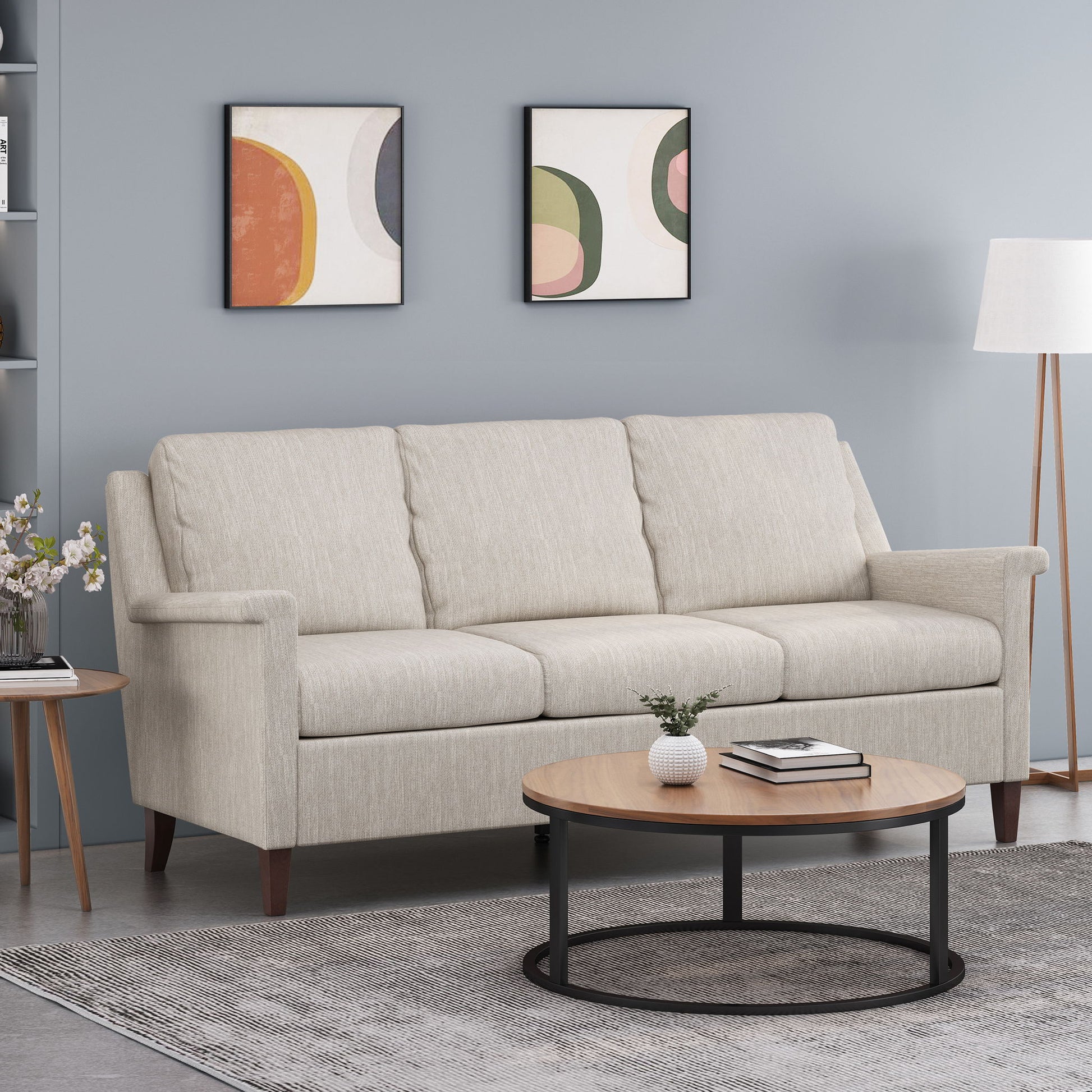 A modern living room featuring a 3 Seater Contemporary Design Sofa in light beige, round wooden coffee table, gray rug, floor lamp, side table with flowers, and two abstract art prints on a light blue wall.