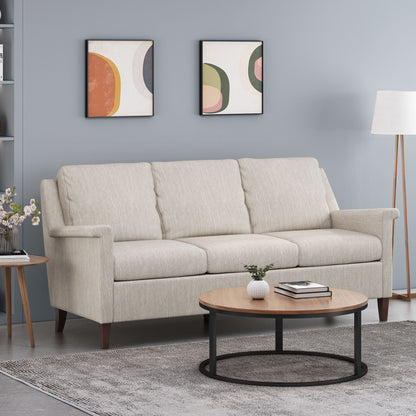 A modern living room featuring a 3 Seater Contemporary Design Sofa in light beige, round wooden coffee table, gray rug, floor lamp, side table with flowers, and two abstract art prints on a light blue wall.