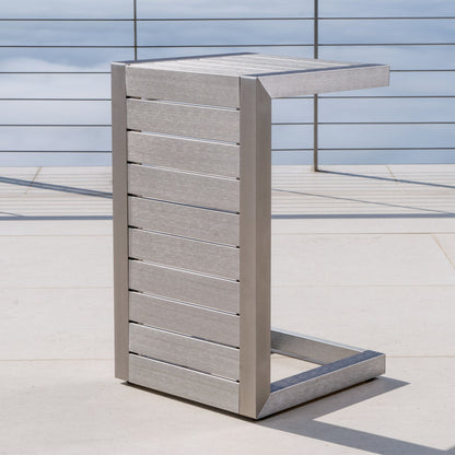 The Aluminum Outdoor C-Shaped Side Table for Patio, featuring a modern silver slatted design and metal frame, is placed on a light patio floor with metal railings and a cloudy sky in the background.