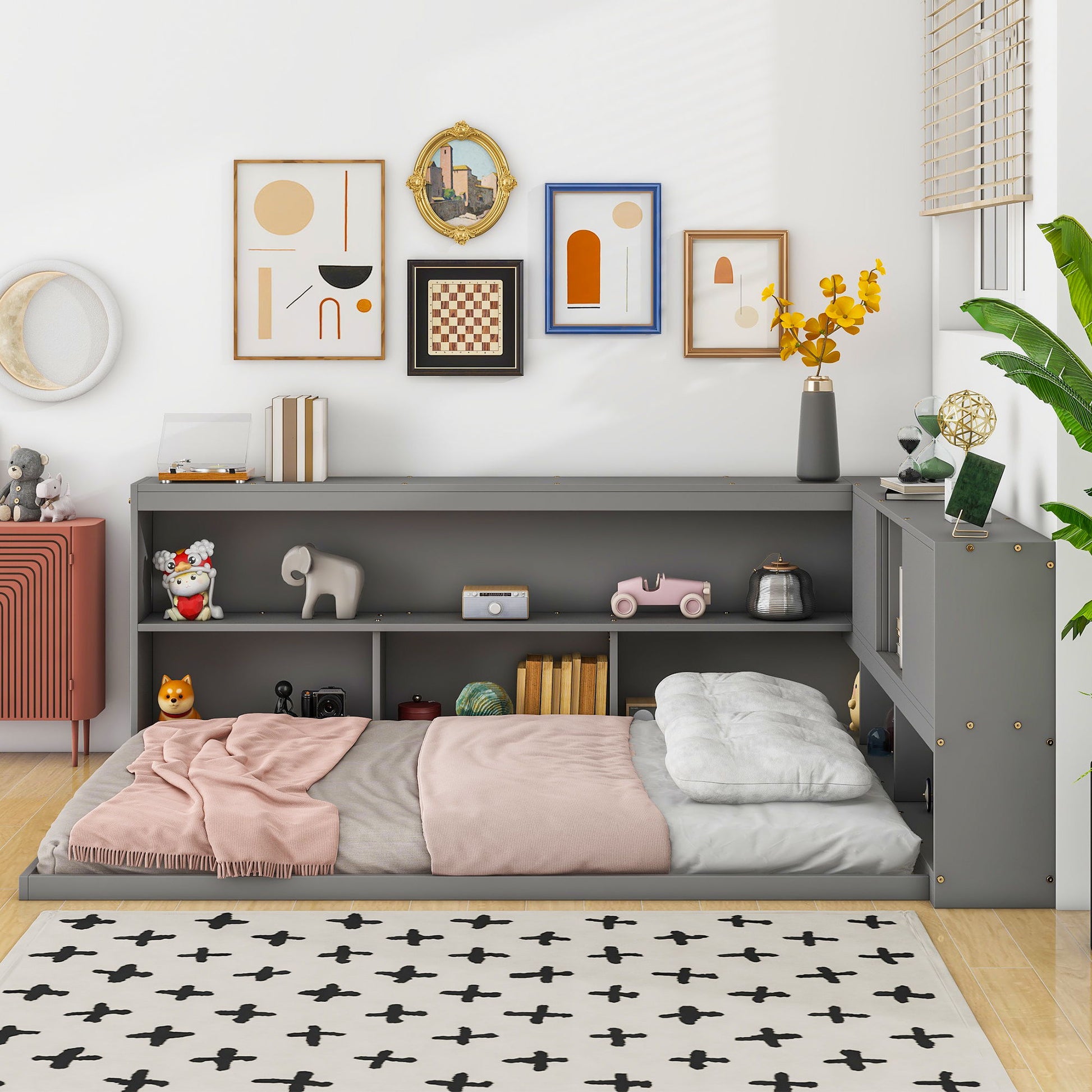 A cozy kids' bedroom featuring the Floor Bed With L-Shaped Bookcases, Sliding Doors, Without Slats in gray, pink bedding, toy- and book-filled shelves, framed art, a white rug with black crosses, a red cabinet, and a potted plant.