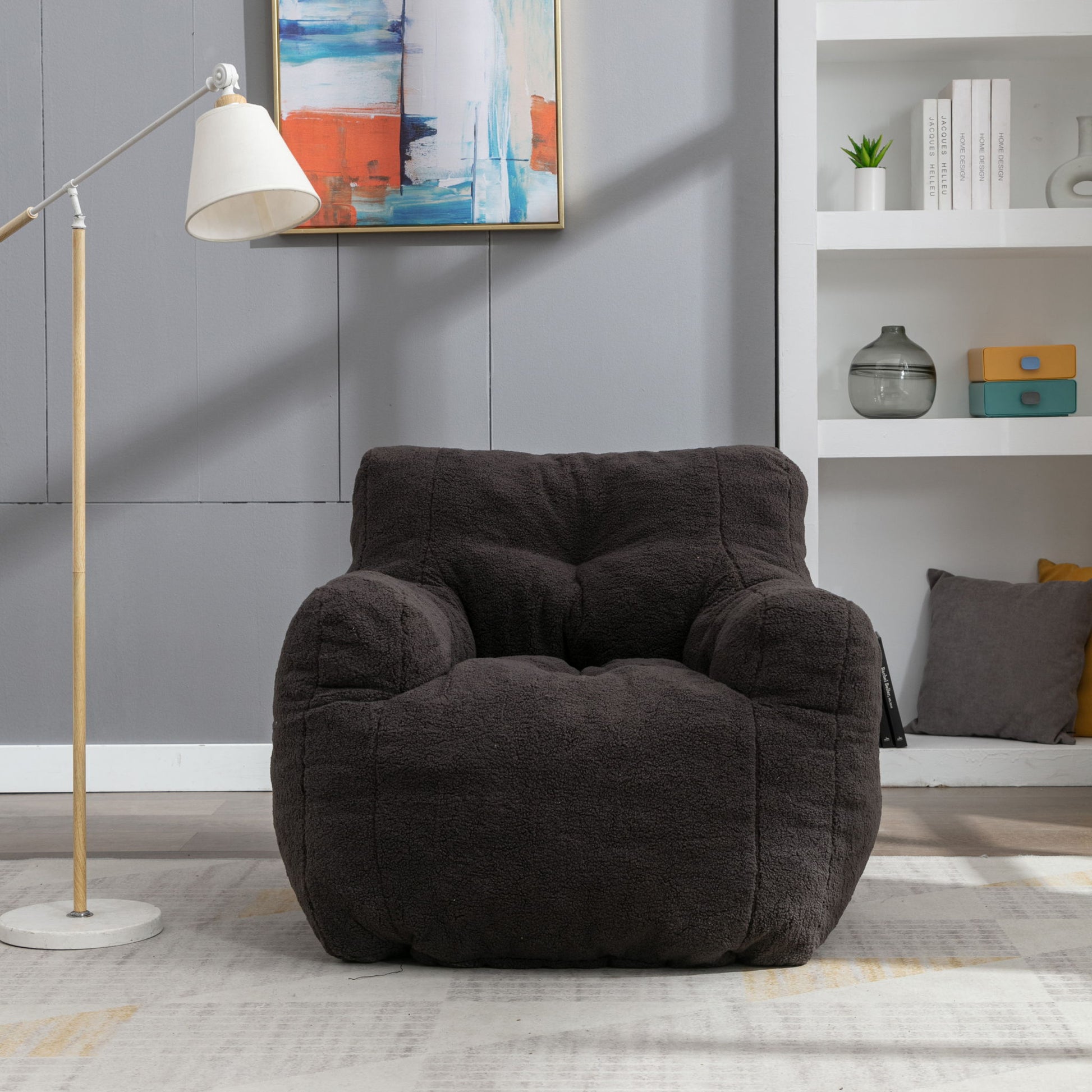 A Soft Teddy Fabric Tufted Foam Bean Bag Chair sits on a light rug in a modern living room with gray walls, a floor lamp, colorful abstract art, and white shelves holding books and decor.