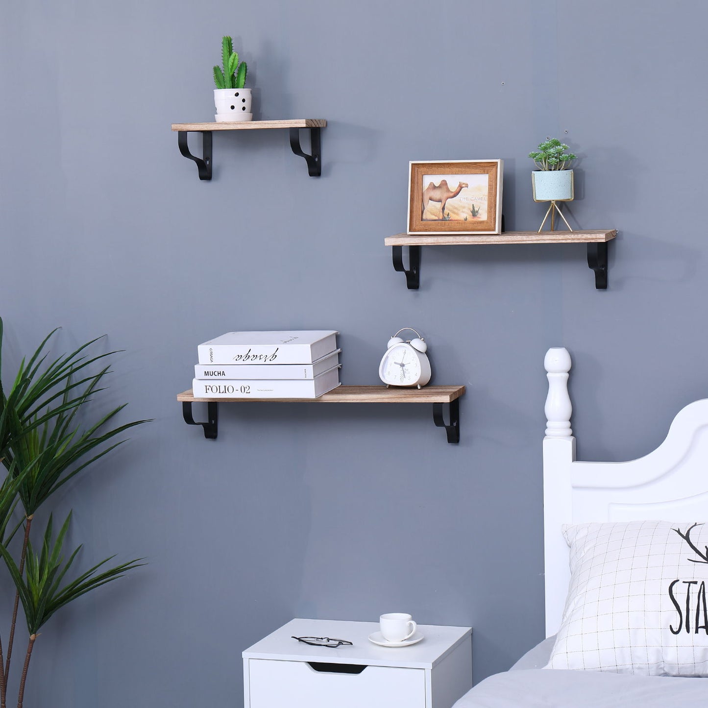 A set of three Long Wall Shelves in natural wood is wall-mounted above a bed and nightstand, displaying a potted cactus, framed picture, alarm clock, books, and plant. These Floating Shelves add stylish storage to the bedroom.