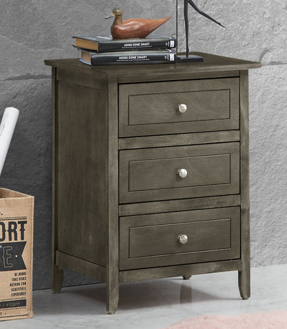 The Daniel 3 Drawer Nightstand with silver knobs stands against a gray wall. On top are three stacked books, a wooden duck figurine, and part of a black metal sculpture. A burlap bag is partially visible on the left.