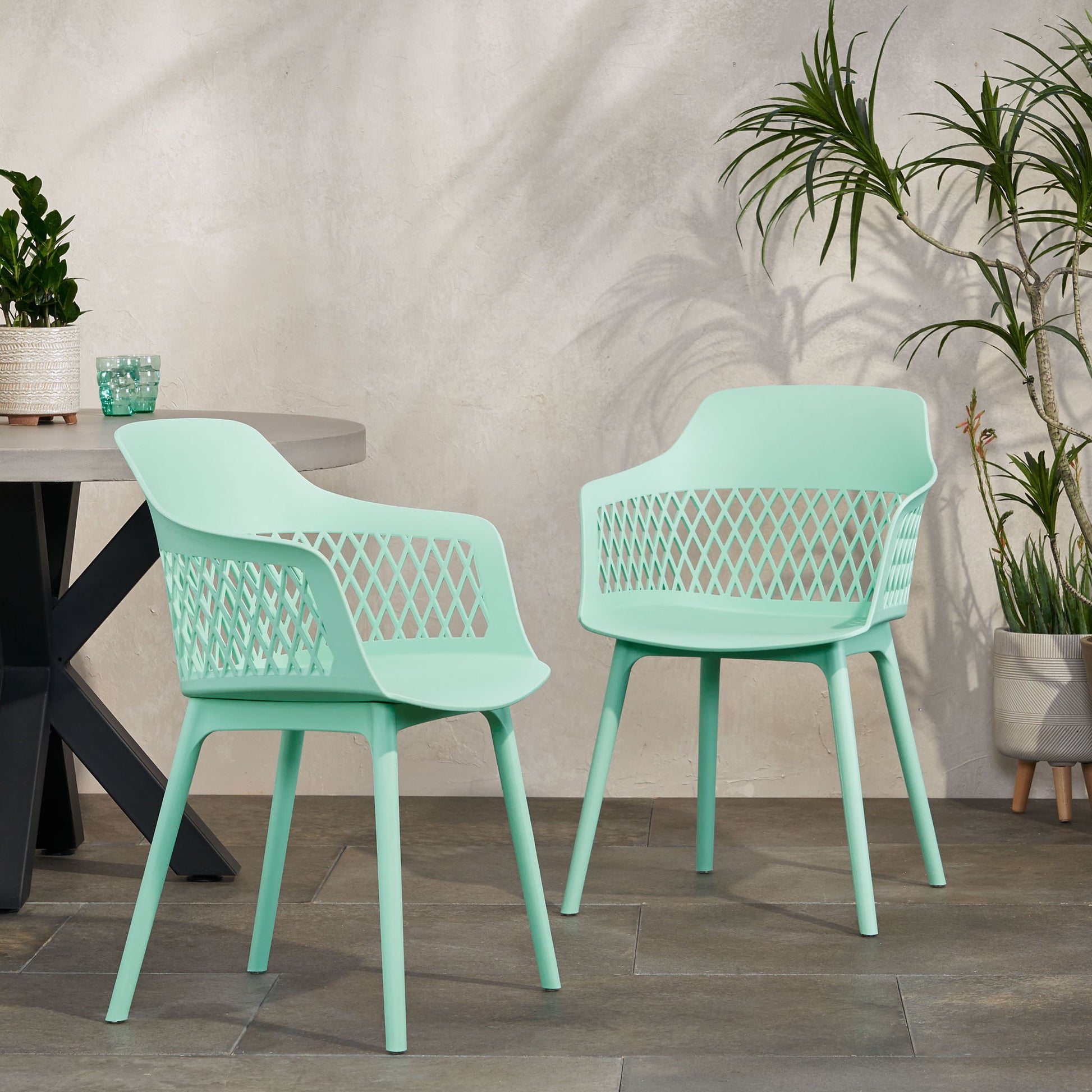 Two Azalea chairs (set of 2) in light green modern plastic with perforated backs are placed on a tiled patio near a round table with potted plants and tall greenery by a light-colored wall.