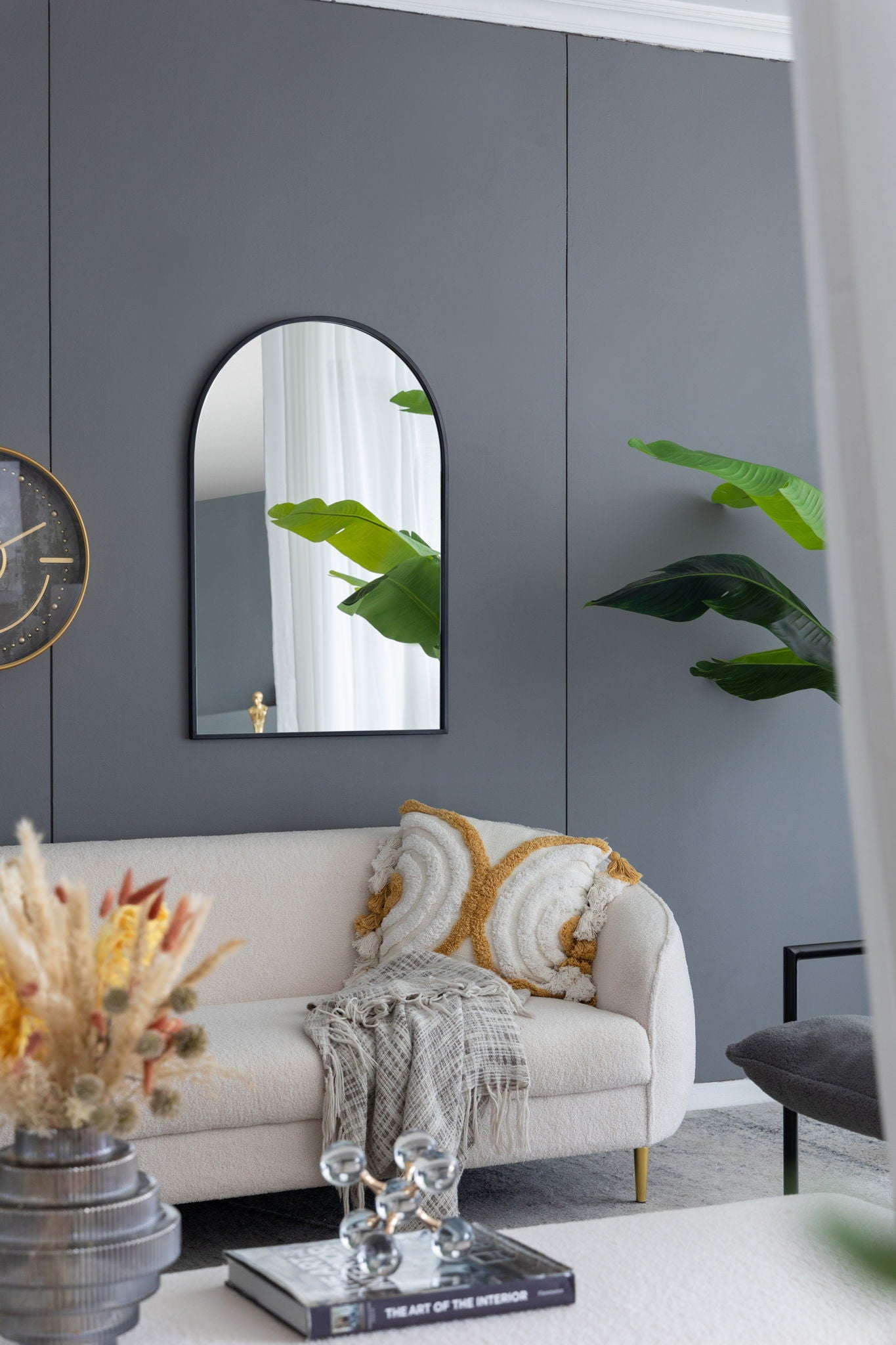 A modern living room with a gray wall, white sofa, yellow and white pillows, patterned throw, round clock, Arched Mirror With Metal Frame, potted plants, and a coffee table with books, glass decor, and dried flowers.