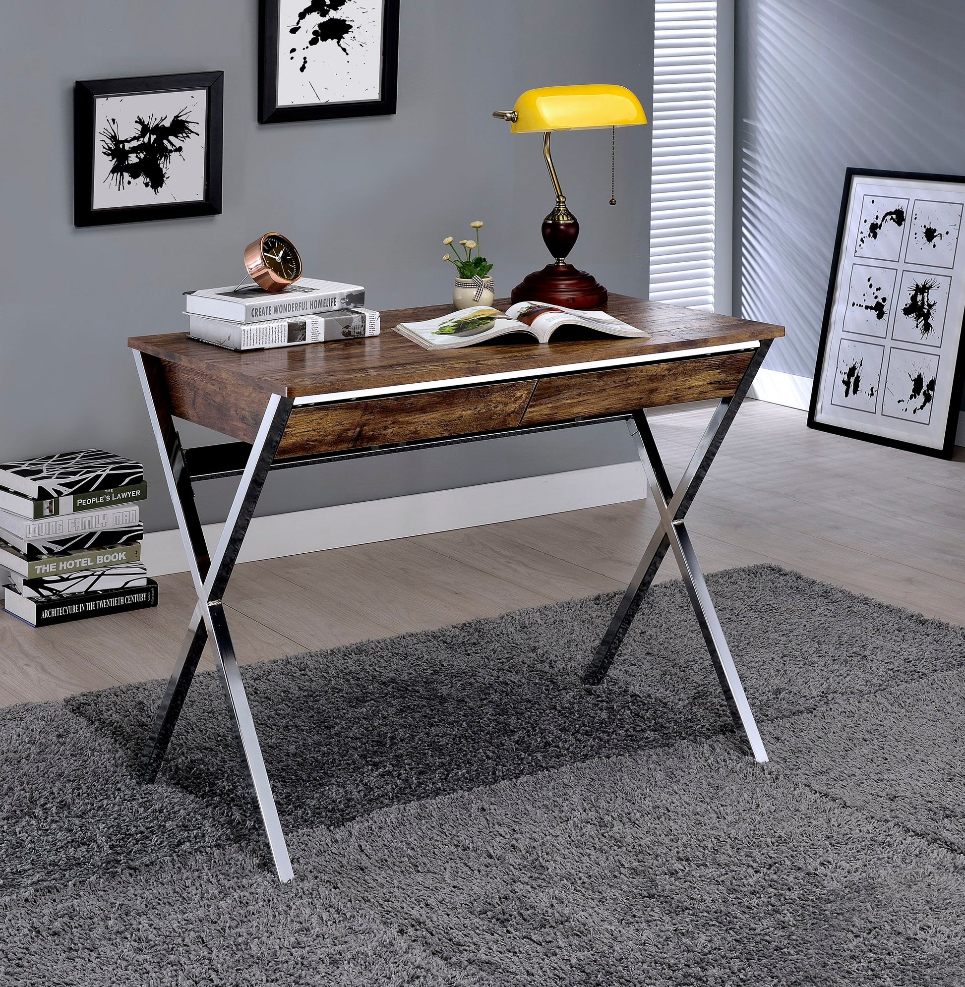 The Callers Weathered Writing Desk in Chrome and Oak features on a gray rug with an open book, potted plant, golden lamp, and decorative clock on top. Abstract wall art and a stack of books complete the modern room decor.