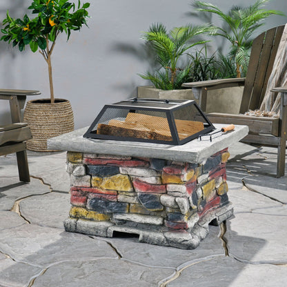 The Outdoor Wood Burning Magnesium Oxide Fire Pit with Lid, featuring colorful stacked stones and a metal mesh cover, sits on a stone patio surrounded by wooden chairs and green potted plants.