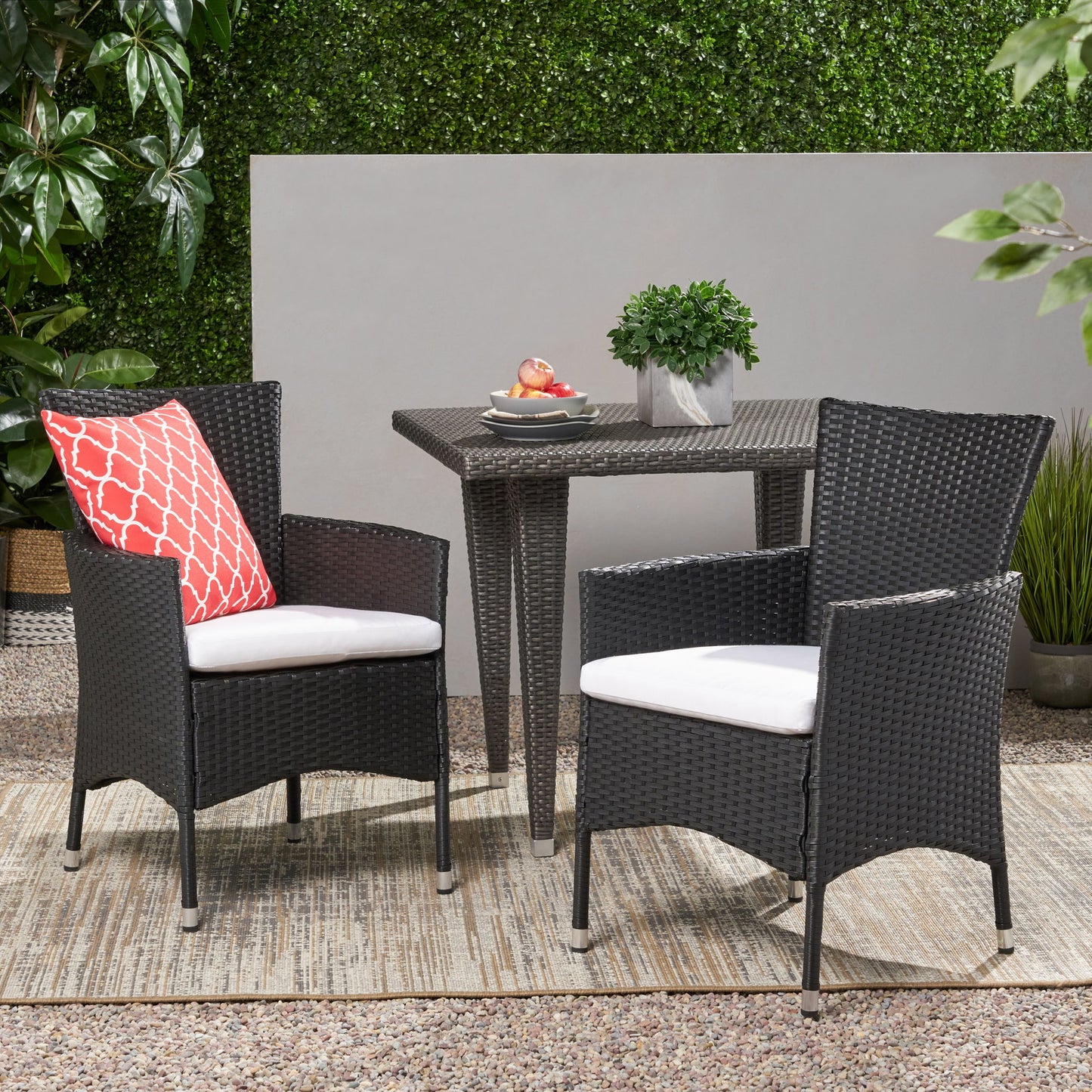 Two Dimensional Outdoor Dining Chairs with cushion seats and iron frames (set of 2) sit by a square wicker table with a plant and fruit bowl, arranged on a rug with greenery and plants in the background.