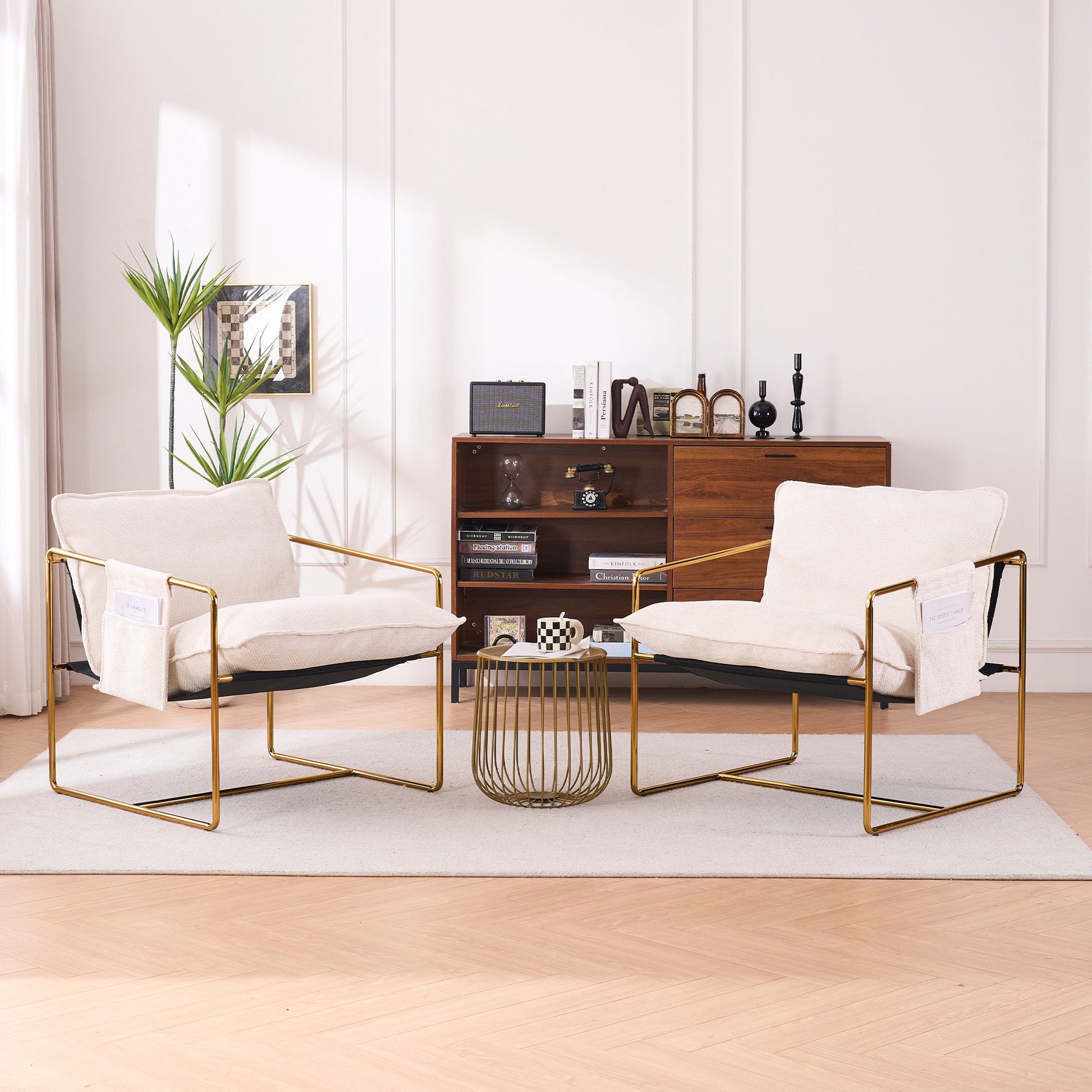 A pair of Upholstered Hanging Armchairs with gold-plated metal frames, arm pockets, and crushed foam cushions in skin-friendly woven fabric face each other across a wire-frame side table in a stylish living room with decor and a tall plant.