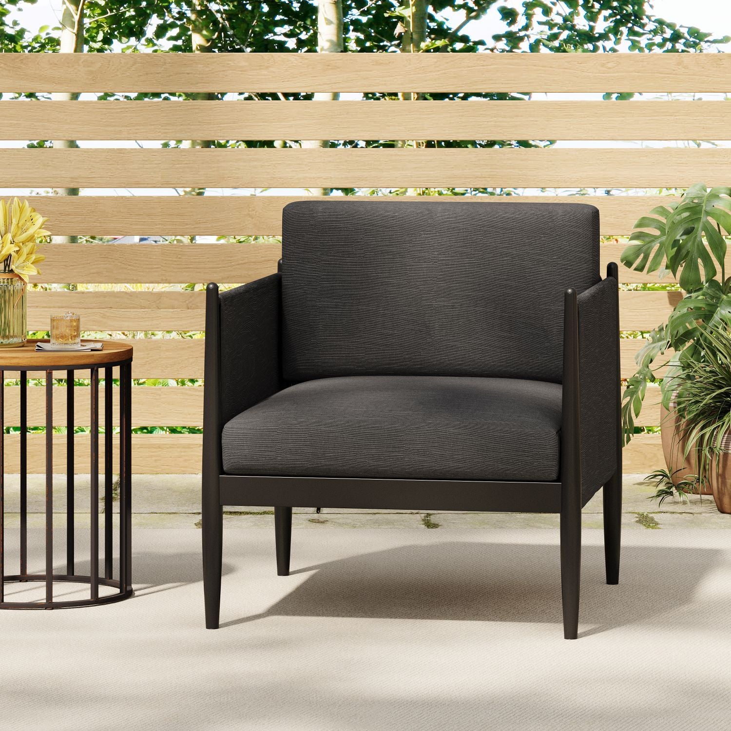 The Dawson - Club Chair, a modern black armchair with square cushions, is placed on a light rug outdoors beside a round side table and green plants, framed by a wooden slat fence and trees in the background.