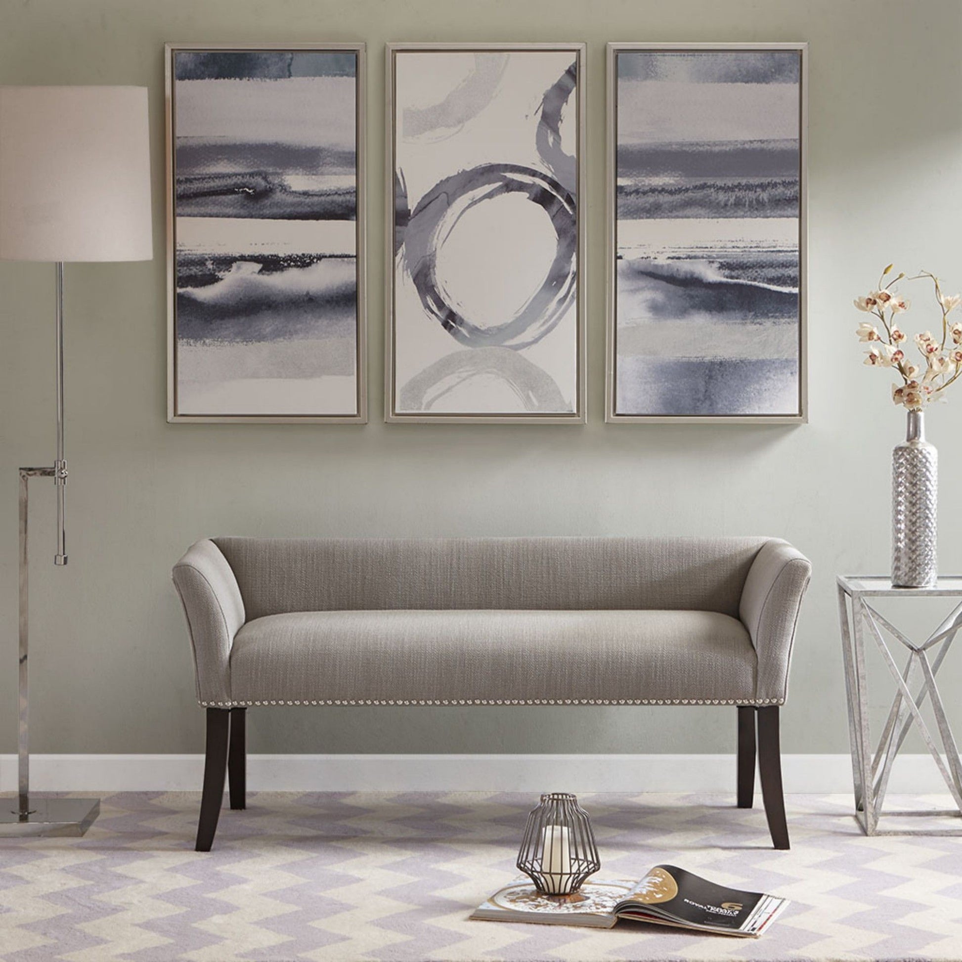 A modern living room featuring the Elegant Design Bench Upholstery with a solid wood frame, three abstract paintings, a floor lamp, glass side table with a vase, plus a magazine and candle holder on a patterned rug.