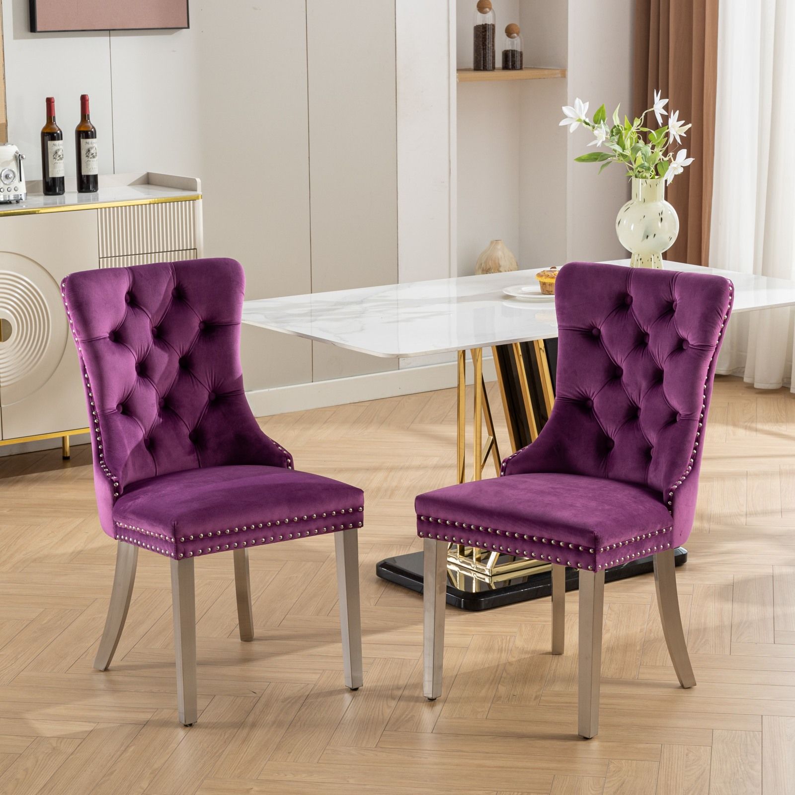 A set of two Nikki modern purple velvet dining chairs with tufted backs, nailhead trim, and chrome stainless steel legs are arranged on a light wood floor near a white marble dining table in an elegant room with decor and wine bottles.