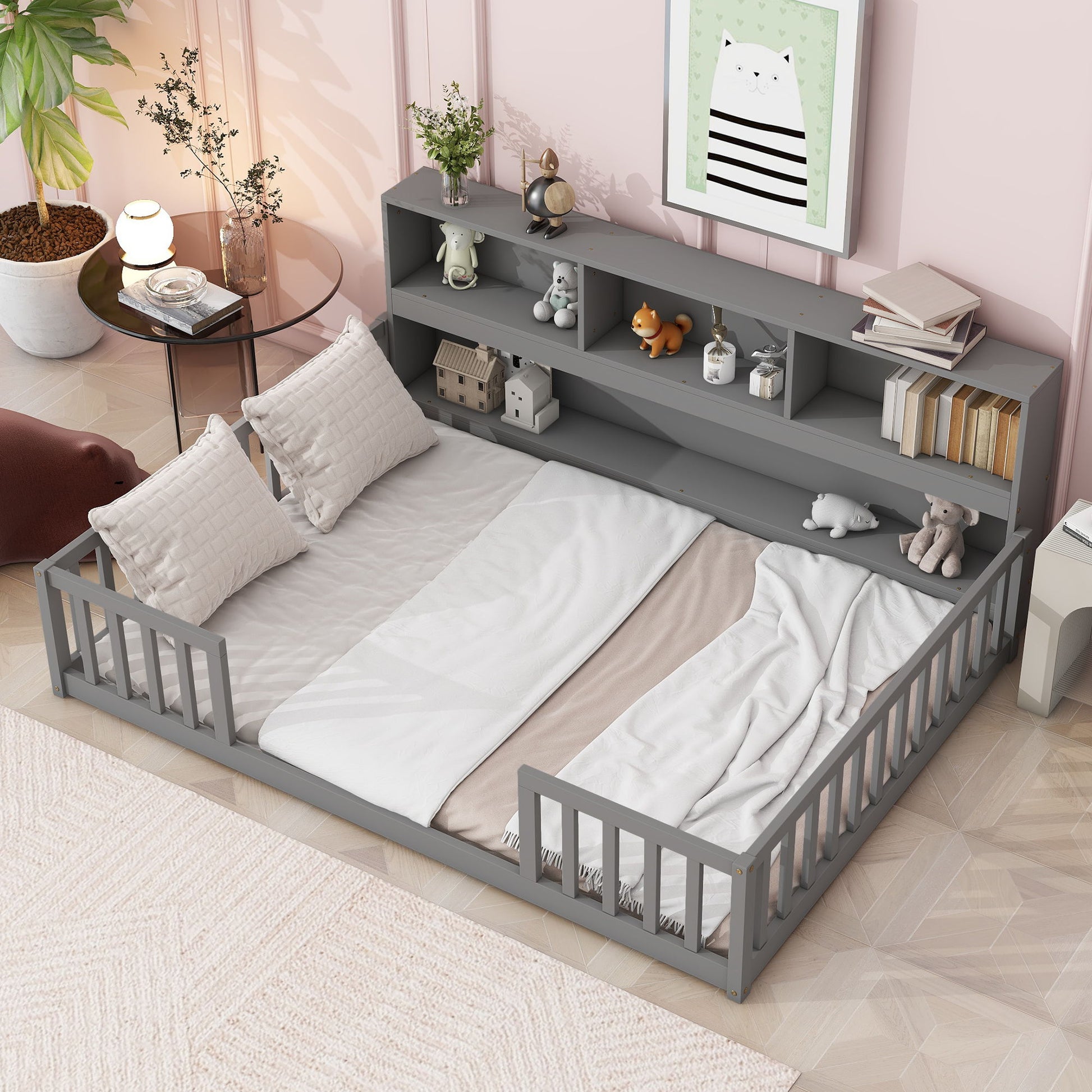 The Floor Bed With Side Bookcase, Shelves, Guardrails features a cozy gray frame, guardrails, white bedding with two pillows and a beige blanket. The headboard shelves display toys and books. Art and plants decorate the softly lit children's room.