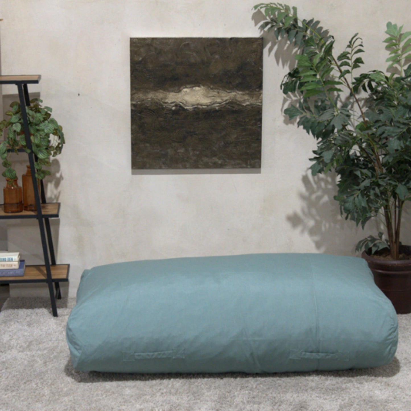 The Kasper - Water Resistant Lounger Bean Bag in blue sits on a carpeted living room floor, with a potted plant, books and plants on shelves, and textured wall art in the background.