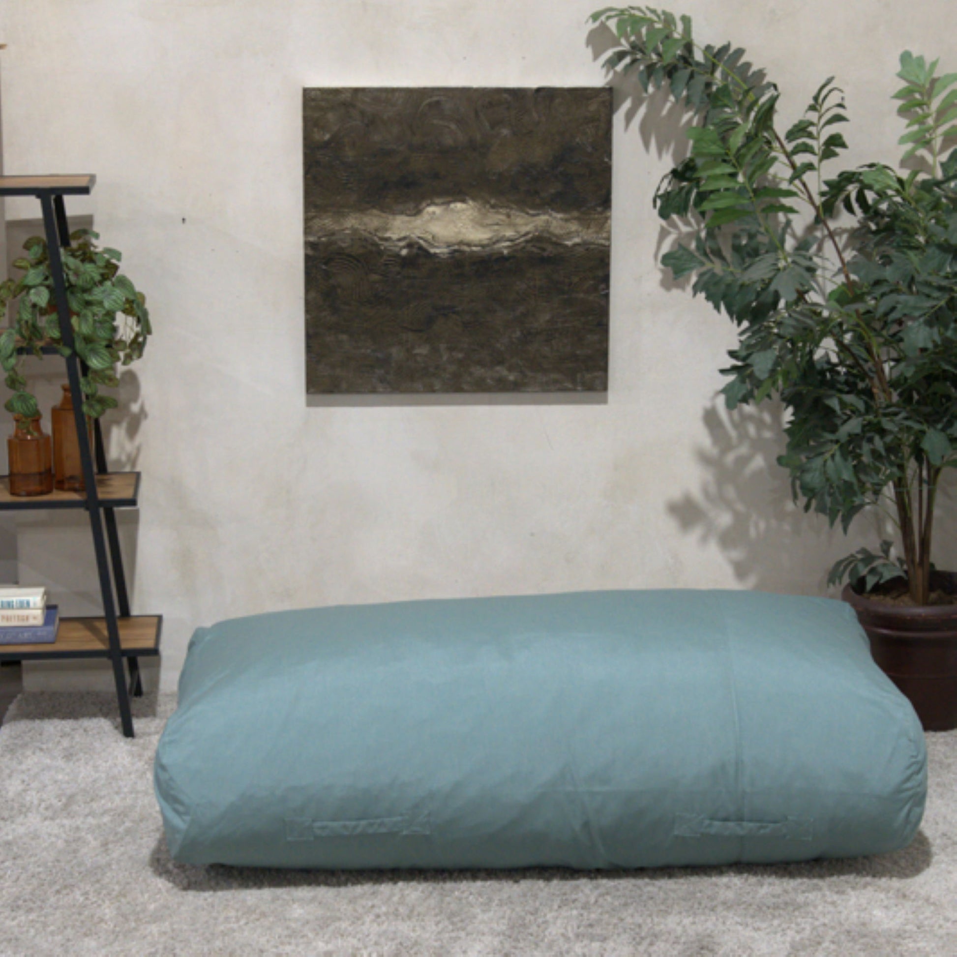 The Kasper - Water Resistant Lounger Bean Bag in blue sits on a carpeted living room floor, with a potted plant, books and plants on shelves, and textured wall art in the background.