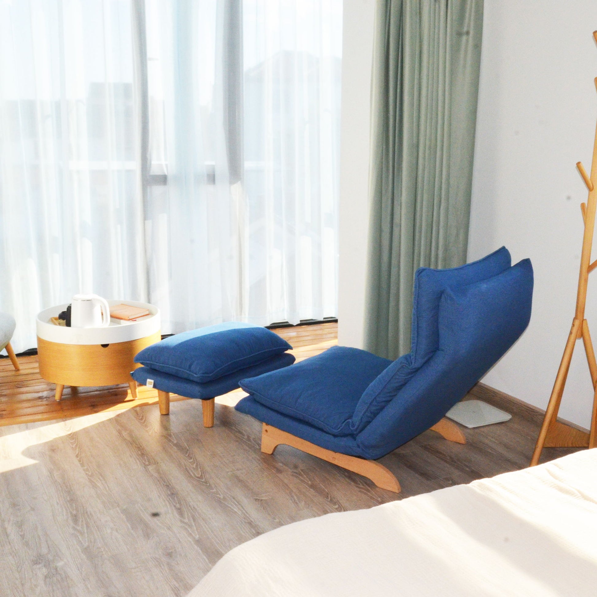 A cozy reading nook features the Single Lazy Sofa Chair Leisure Foldable Reclining Chair in blue with a matching ottoman on wooden floors, beside a small round table with a teapot, by large windows with sheer and green curtains.