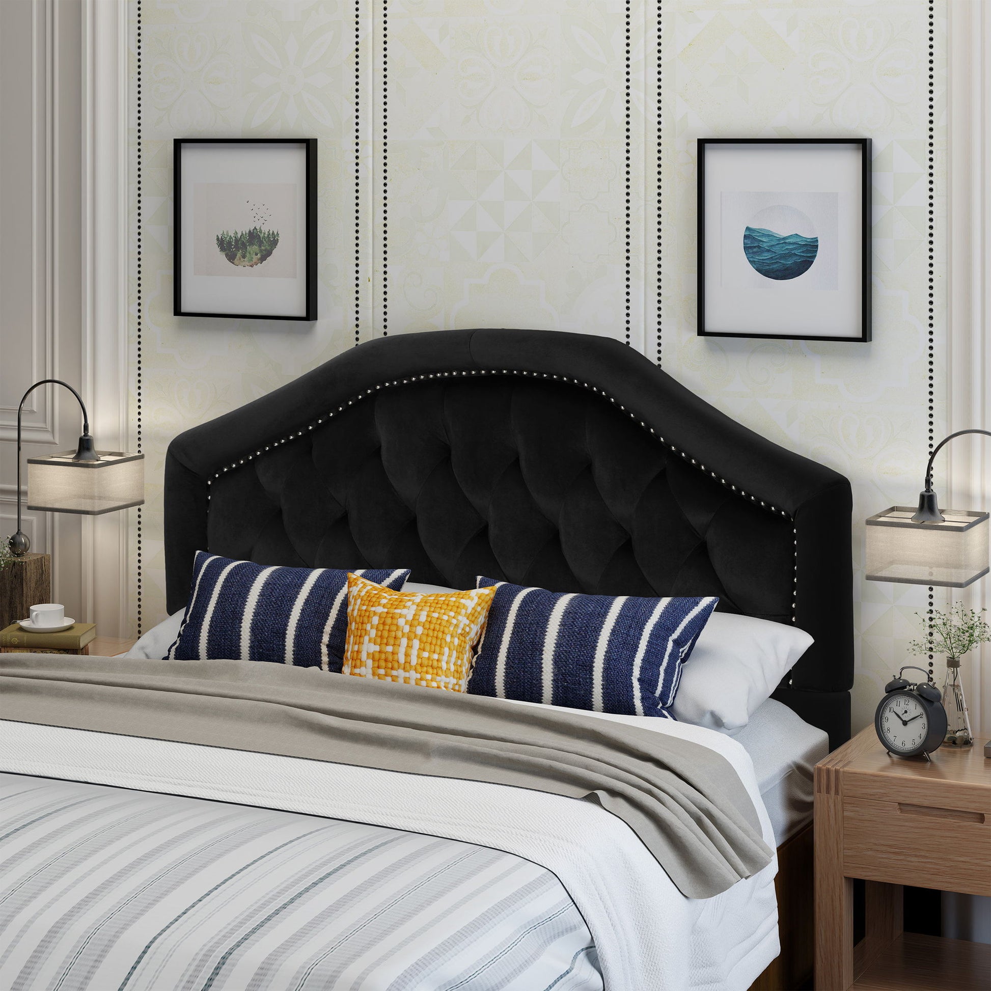 A modern bedroom featuring the Kaarina Contemporary Tufted Fabric Headboard in black, striped bedding, patterned throw pillows, two framed wall pictures, and matching wooden nightstands with lamps, books, a clock, and small plants.