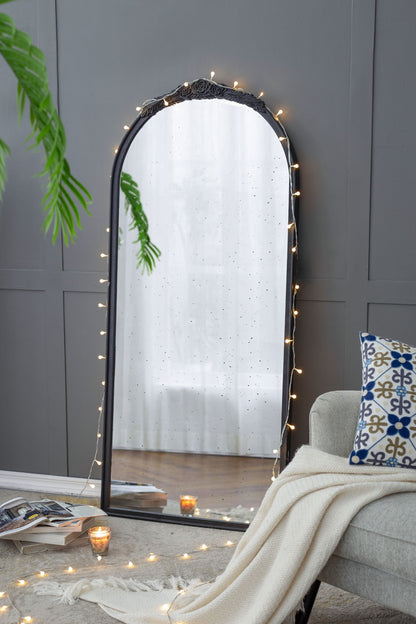 A Hand Carved Rose Antique Mirror Frame, Wood Arch Mirror Wall Decor for Living Room leans against a gray paneled wall, accented with string lights. Nearby sit a cozy chair, patterned cushion, white blanket, open magazine, and glowing candle.