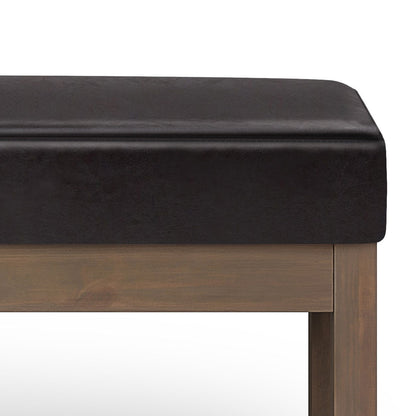 A close-up of the Milltown - Upholstered Ottoman Bench shows its dark brown cushioned seat and wooden frame with straight legs, set against a white background.