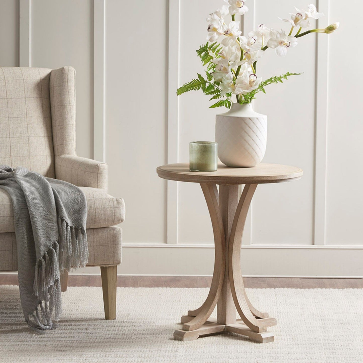 A rustic ash veneer round accent table in reclaimed wheat sits next to a beige armchair with a gray throw, topped with a green candle and white orchid vase, on a light rug before a white paneled wall.