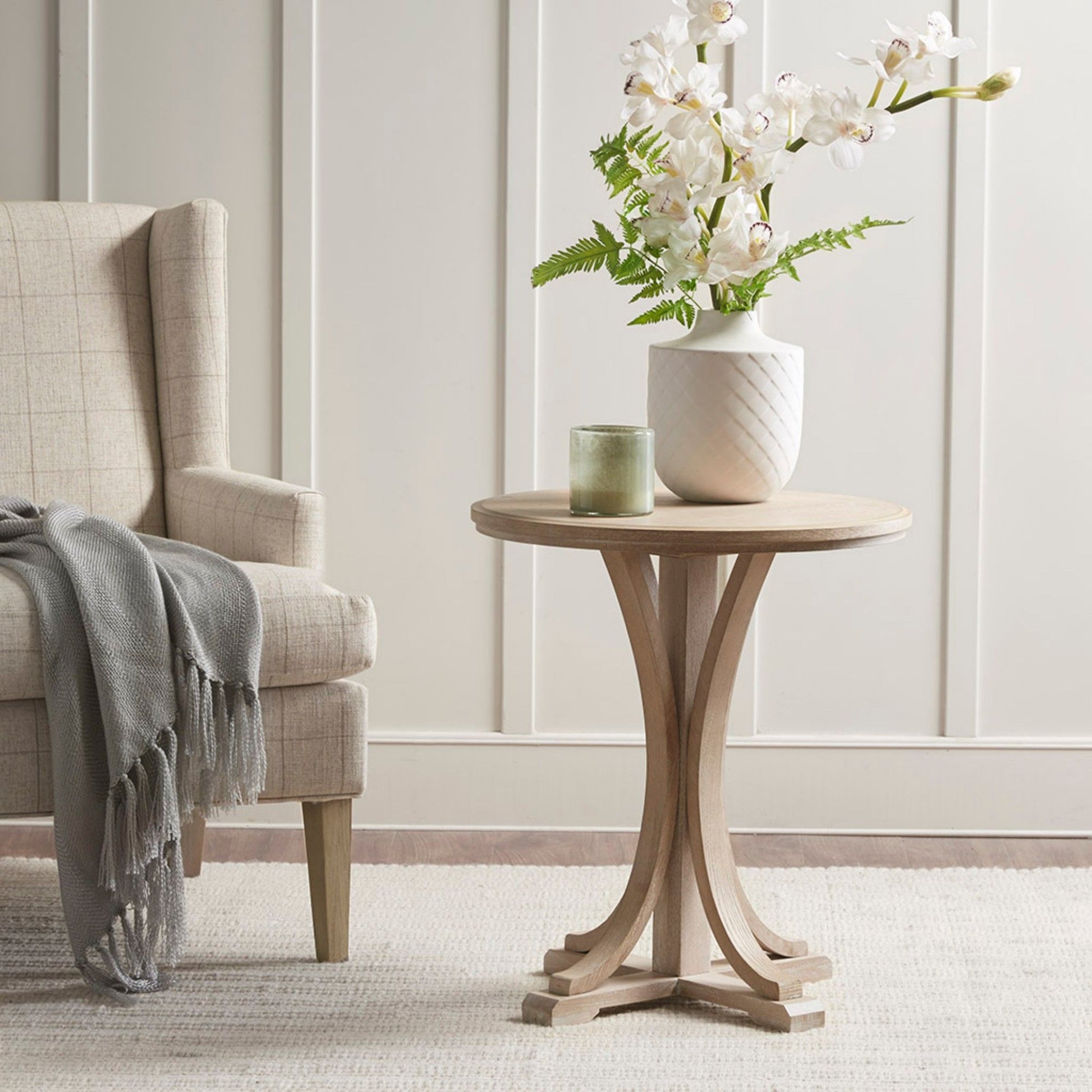 A rustic ash veneer round accent table in reclaimed wheat sits next to a beige armchair with a gray throw, topped with a green candle and white orchid vase, on a light rug before a white paneled wall.