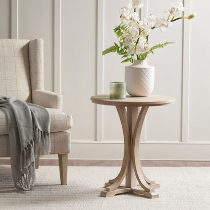A rustic ash veneer round accent table in reclaimed wheat sits next to a beige armchair with a gray throw, topped with a green candle and white orchid vase, on a light rug before a white paneled wall.