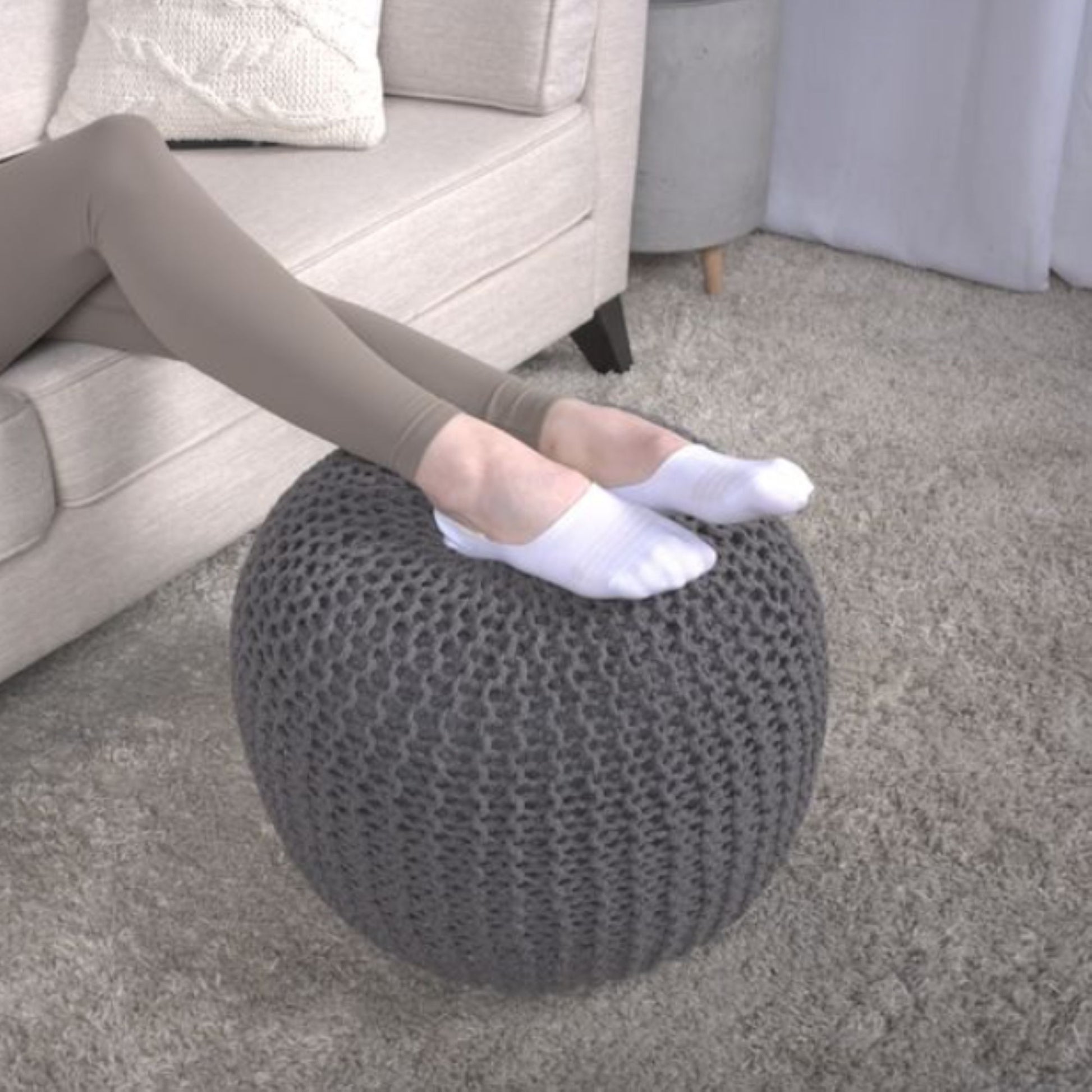 A person in white socks rests their feet on the Bordeaux Knitted Cotton Round Pouf, a versatile ottoman and floor cushion, in a living room with a beige sofa, light carpet, and cushion in the background.