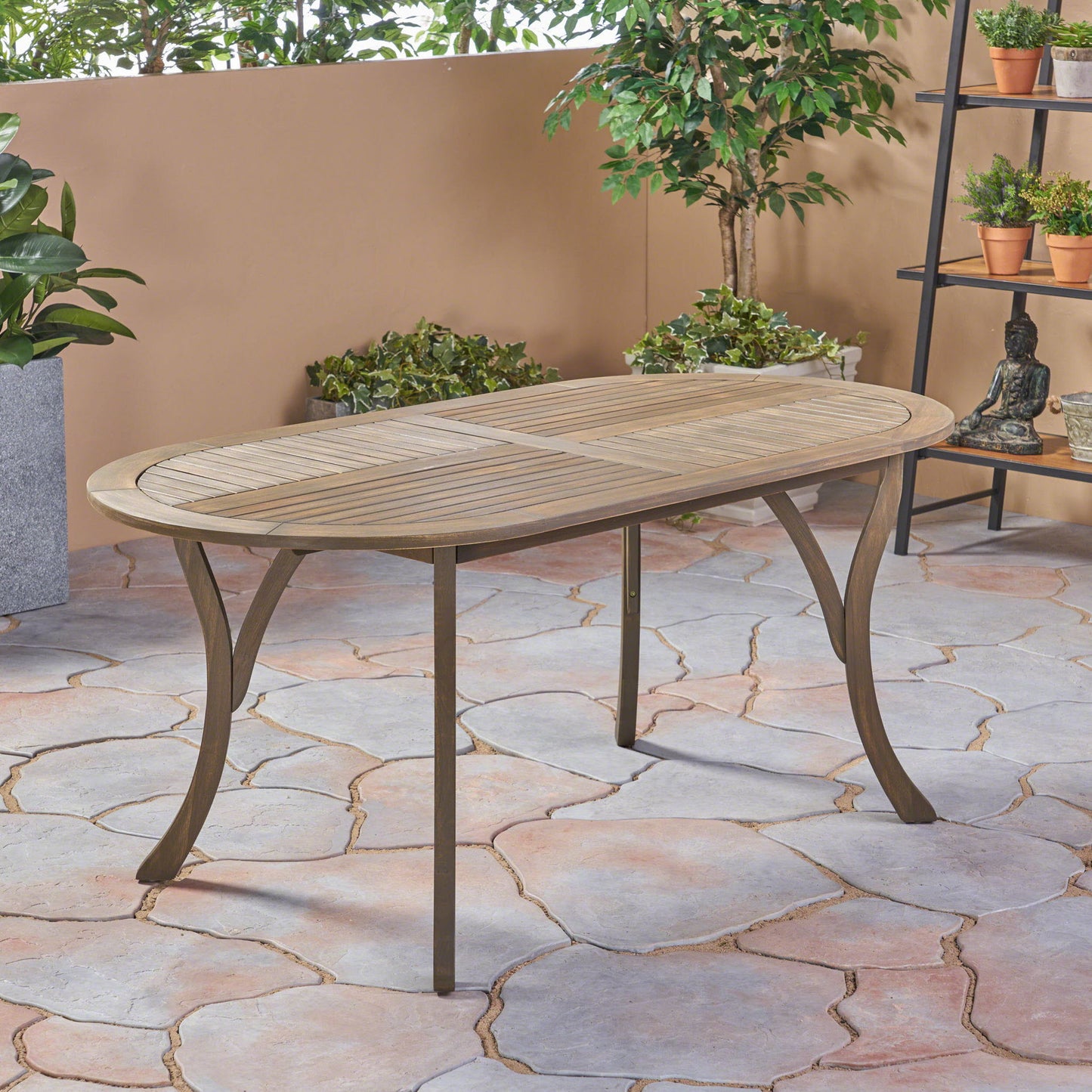 The Outdoor Table With Smooth Lines, Slat Top features an elegant oval wooden design with metal legs, set on a stone-tiled floor amid potted plants and greenery by a beige wall in an inviting outdoor space.