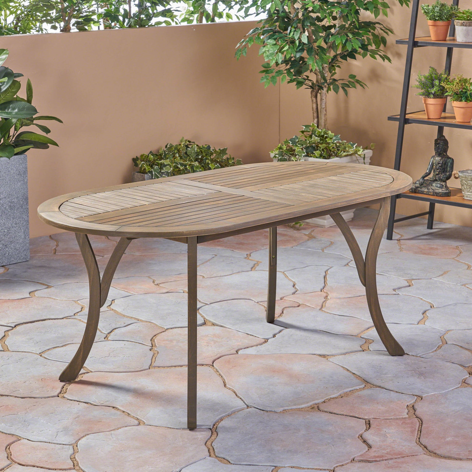 The Outdoor Table With Smooth Lines, Slat Top features an elegant oval wooden design with metal legs, set on a stone-tiled floor amid potted plants and greenery by a beige wall in an inviting outdoor space.