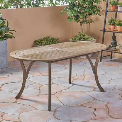 The Outdoor Table With Smooth Lines, Slat Top features an elegant oval wooden design with metal legs, set on a stone-tiled floor amid potted plants and greenery by a beige wall in an inviting outdoor space.
