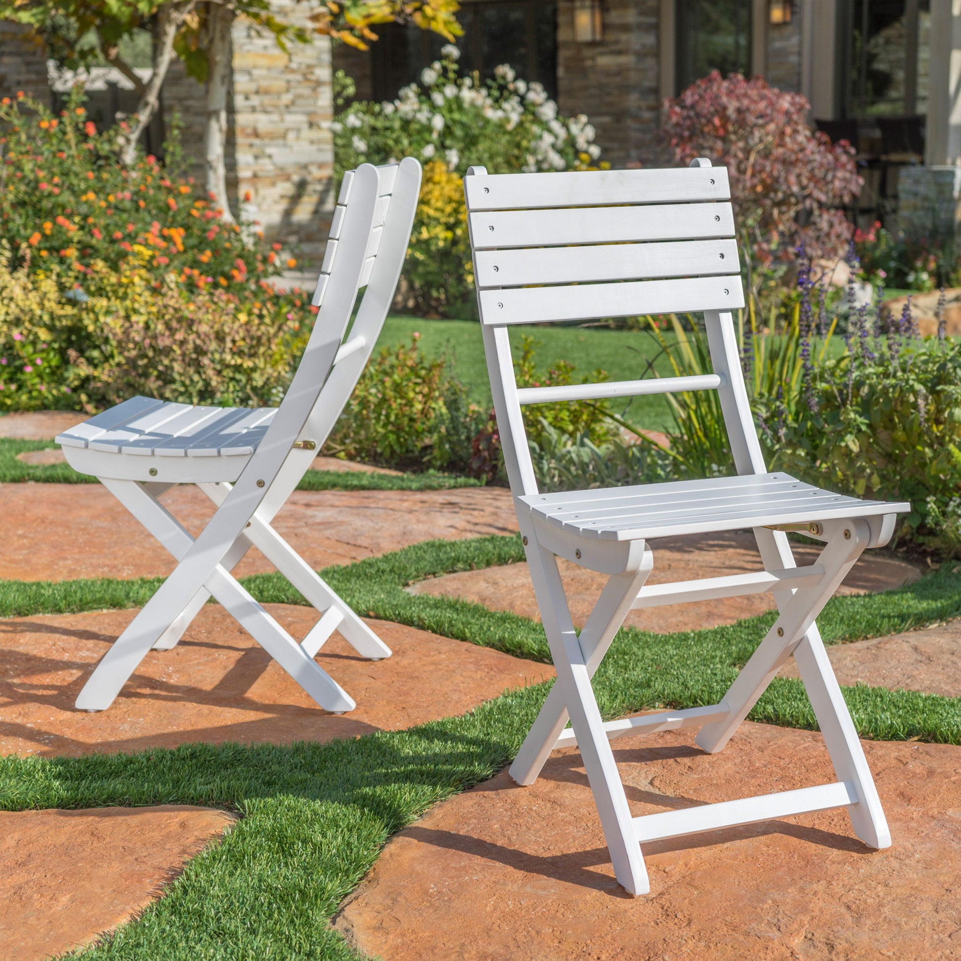 Two Outdoor Foldable Wood Dining Chairs with a slat design are set on a stone patio in a garden, surrounded by green grass and colorful flowers, with a house visible in the background.