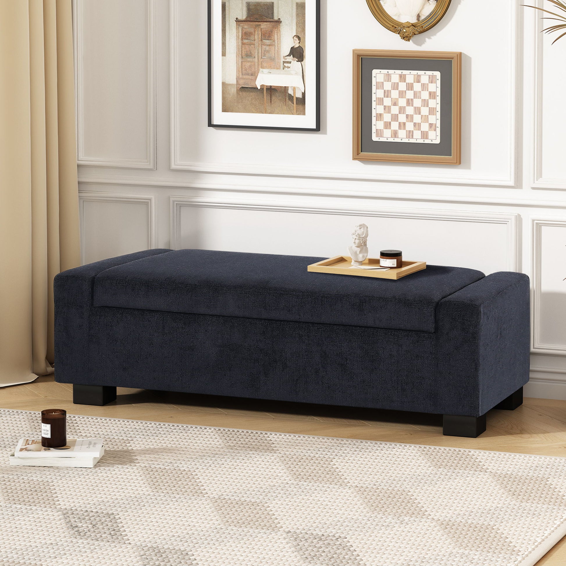 A modern living room featuring a Textured Fabric Ottoman Bench With Hinged Lid in dark blue, topped with a wooden tray of decor, complemented by a patterned rug and framed wall art on white paneled walls.