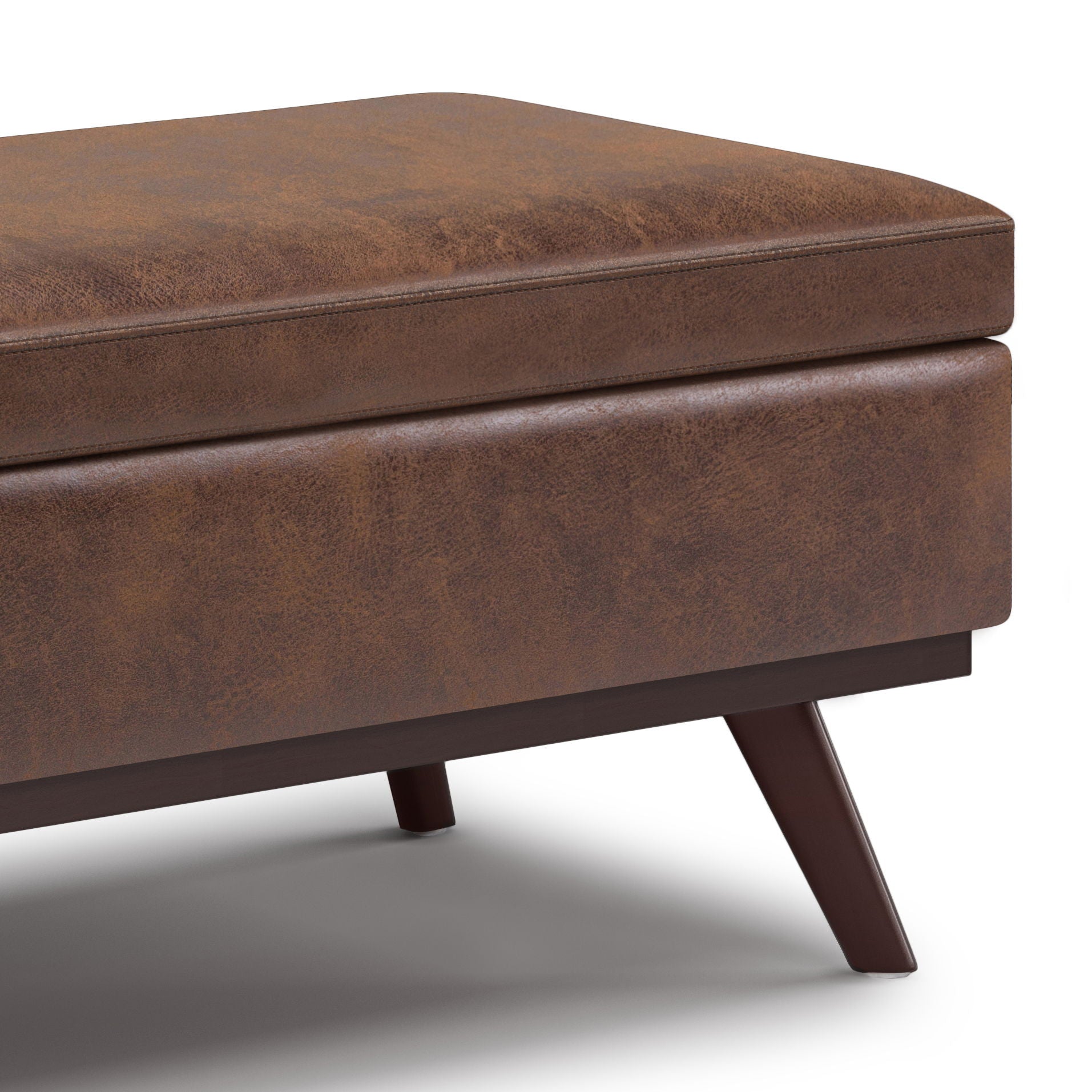 Close-up of the Owen Lift Top Large Coffee Table Storage Ottoman in brown faux leather with angled wooden legs on a white background. The mid-century style upholstery features a subtly textured, worn look.