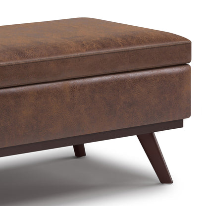 Close-up of the Owen Lift Top Large Coffee Table Storage Ottoman in brown faux leather with angled wooden legs on a white background. The mid-century style upholstery features a subtly textured, worn look.