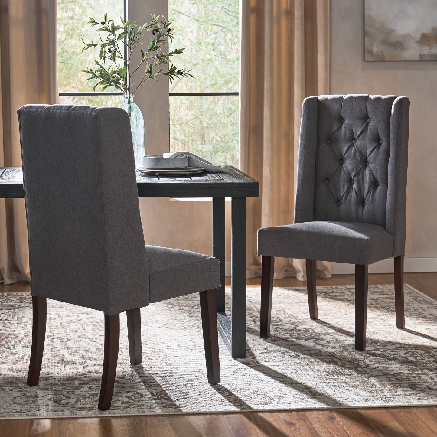 Two Sophisticated Tufted Dining Chairs With High Back Design and dark wooden legs sit by a dark table near a window with light brown curtains. A vase with greenery, a book on the table, and a patterned rug complete the setting.