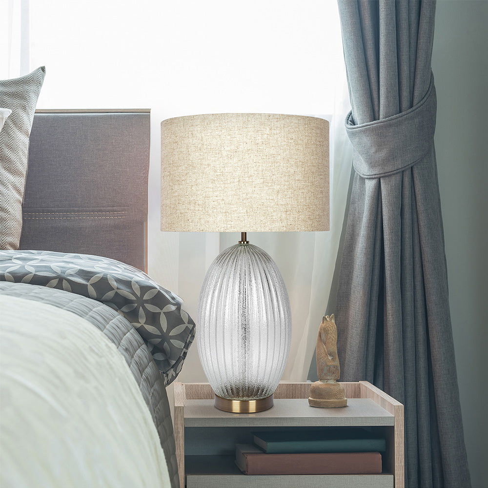 A cozy bedroom features a bed with patterned bedding, a wooden nightstand, and the Aurora Lamp with a ribbed clear base and large beige linen shade. A draped curtain by the window enhances the serene atmosphere.