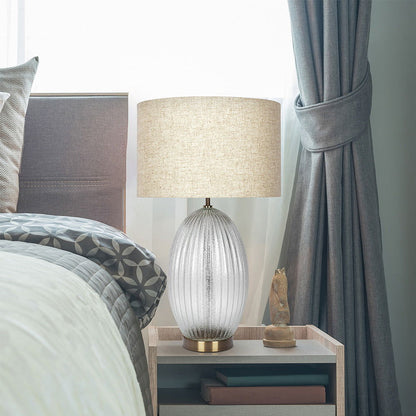 A cozy bedroom features a bed with patterned bedding, a wooden nightstand, and the Aurora Lamp with a ribbed clear base and large beige linen shade. A draped curtain by the window enhances the serene atmosphere.