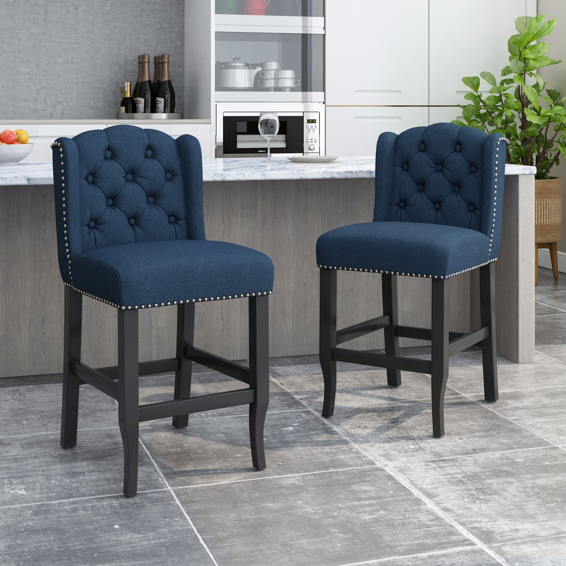 Two Vienna Fabric Tufted Counter Stools with silver nailhead accents (set of 2) are placed before a modern kitchen island with a marble countertop, light wood base, white cabinets, and a potted plant.