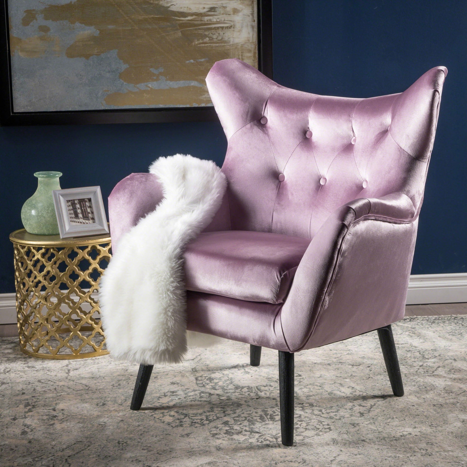 The Alyssa Mid Century Upholstered Arm Chair with wingback design in velvet pink sits on a rug, draped with a white faux fur throw. Beside it, a gold side table holds a green glass vase and framed photo against a blue wall.