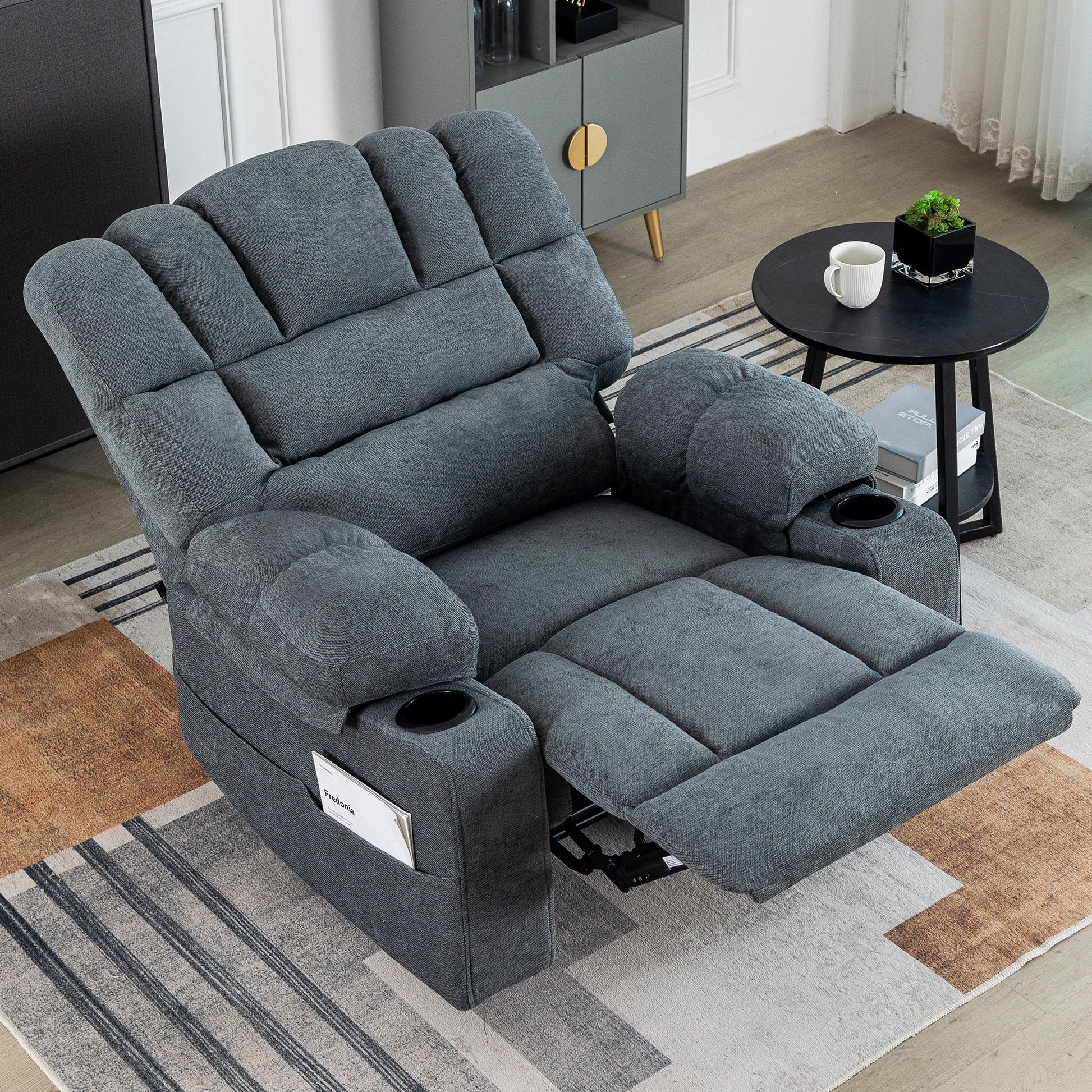 The Massage Recliner Chair Sofa With Heating Vibration, featuring cup holders and a side pocket, sits on a modern area rug in the living room beside a small round black table with a potted plant and white mug.