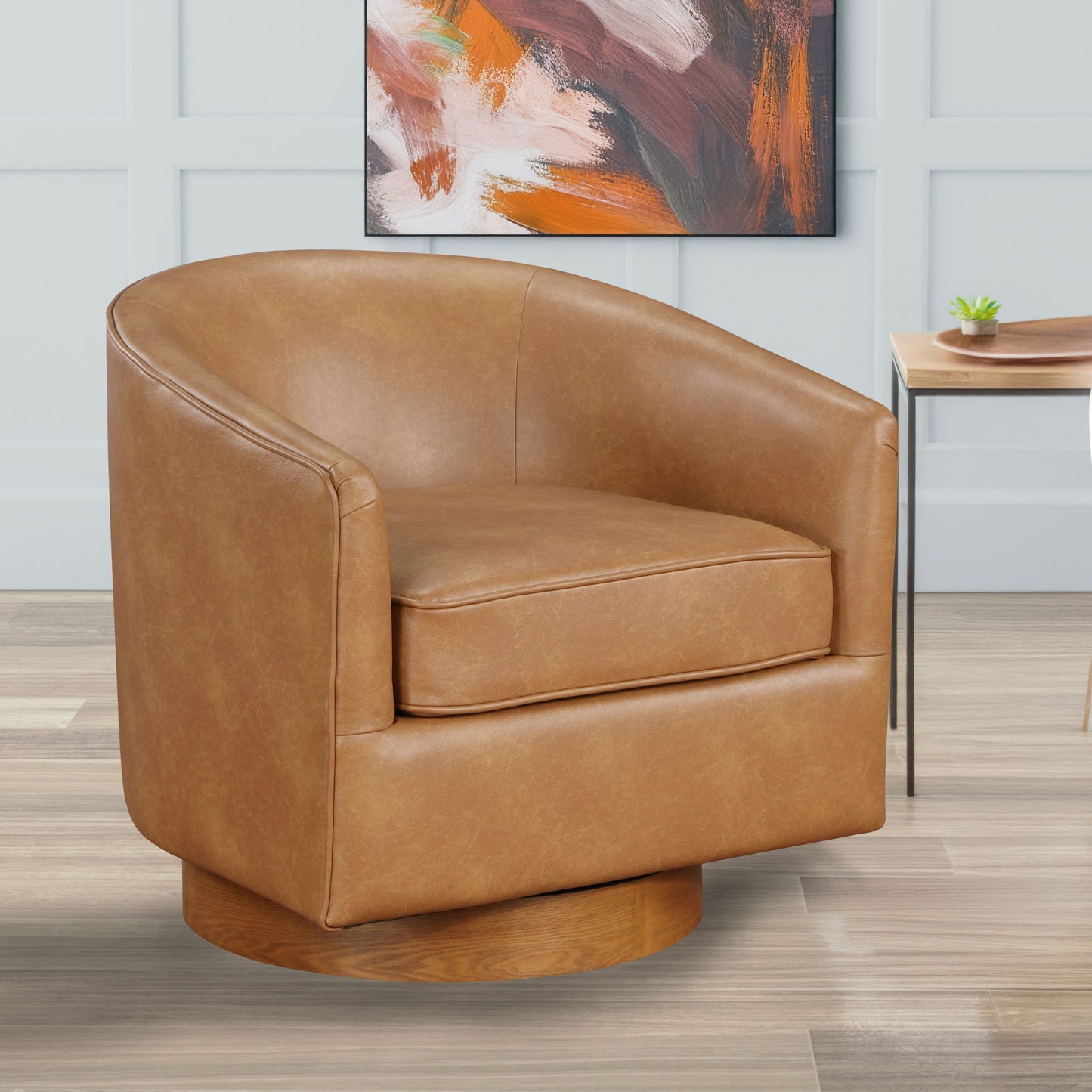 The Maisy - Barrel Swivel Chair in tan leather with a rounded back and wooden base sits on a light wood floor. In the background, there’s a modern abstract painting and a small table topped with a potted plant.
