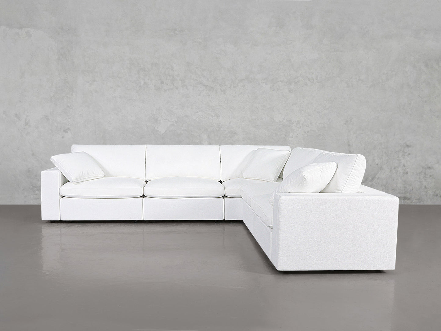 The 5-Seat Modular Corner Sectional features a modern white design with clean lines, plush cushions, and matching throw pillows, placed against a plain gray wall and floor.