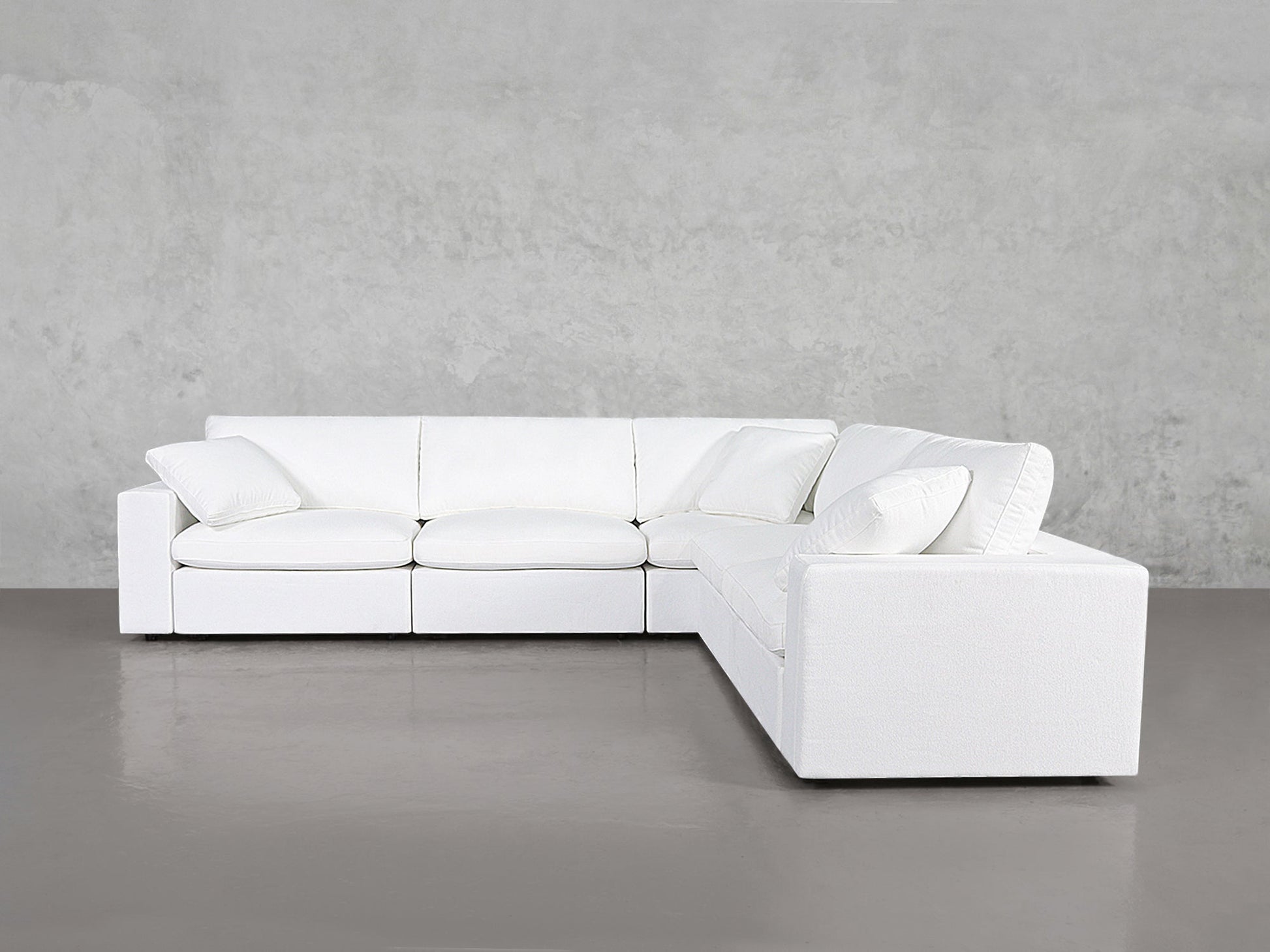 The 5-Seat Modular Corner Sectional features a modern white design with clean lines, plush cushions, and matching throw pillows, placed against a plain gray wall and floor.