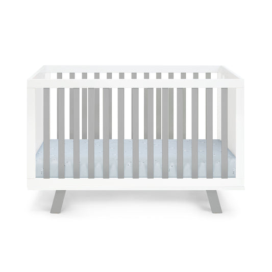 The Livia 3-in-1 Convertible Island Crib features a modern white frame with vertical slats, light grey angled legs, and a light blue mattress, displayed against a plain white background.
