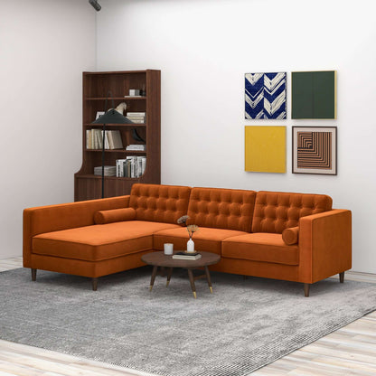 A modern living room featuring the Christian Mid-Century Modern Design Sectional Sofa in orange, paired with a round coffee table, gray rug, wooden bookshelf, and four colorful geometric art pieces on a white wall.