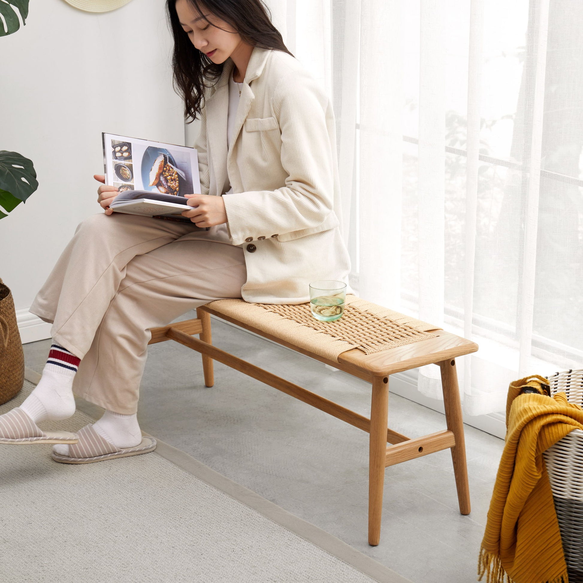 A woman in beige sits on the Woven Design Dining Bench for Dining Room, Bedroom - Natural Oak by a sunlit window, reading a cookbook. A glass of water is beside her and a yellow blanket drapes over the bench’s edge.