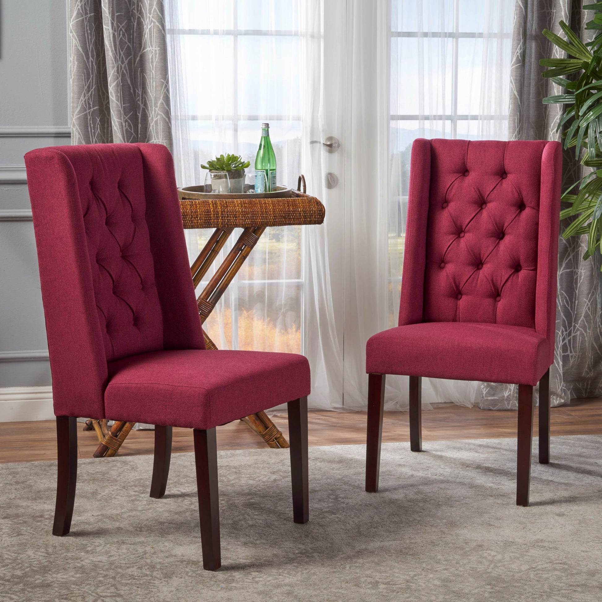 Two Sophisticated Tufted Dining Chairs With High Back Design, upholstered in red and featuring dark wooden legs, sit atop a gray rug in a bright room with large windows, sheer curtains, a wicker table, and a potted plant in the background.