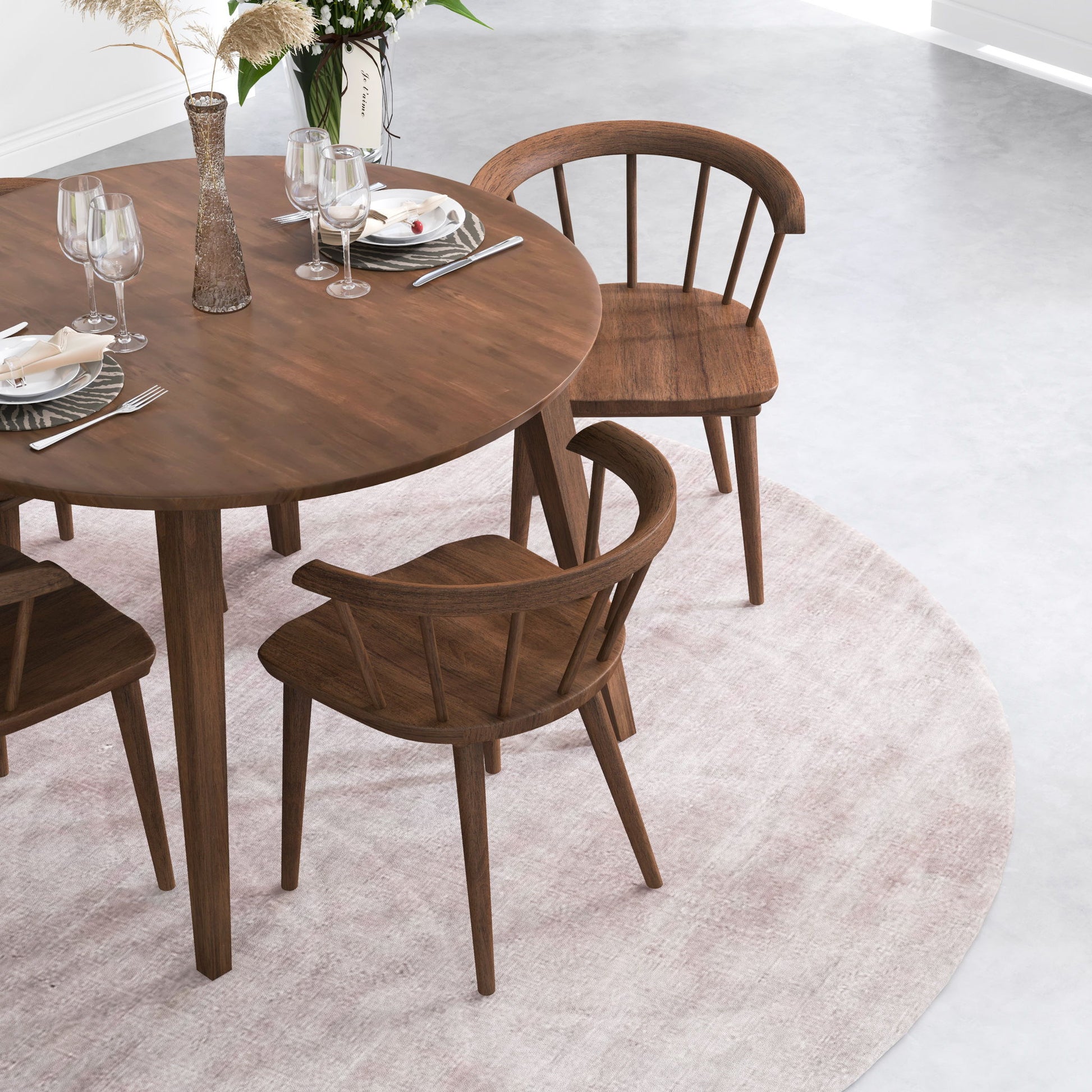 A round wooden dining table set with plates, glasses, and cutlery is surrounded by Kingsley Dining Chairs (Set of 2) in brown on a light pink circular rug in a modern, bright room.